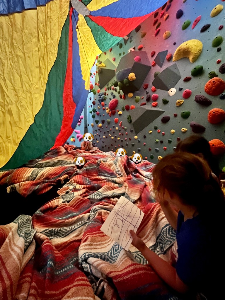 Inside of DIY tent made from giant parachute inside youth bouldering gym area, with 10+ children lying with yoga blankets inside of tent